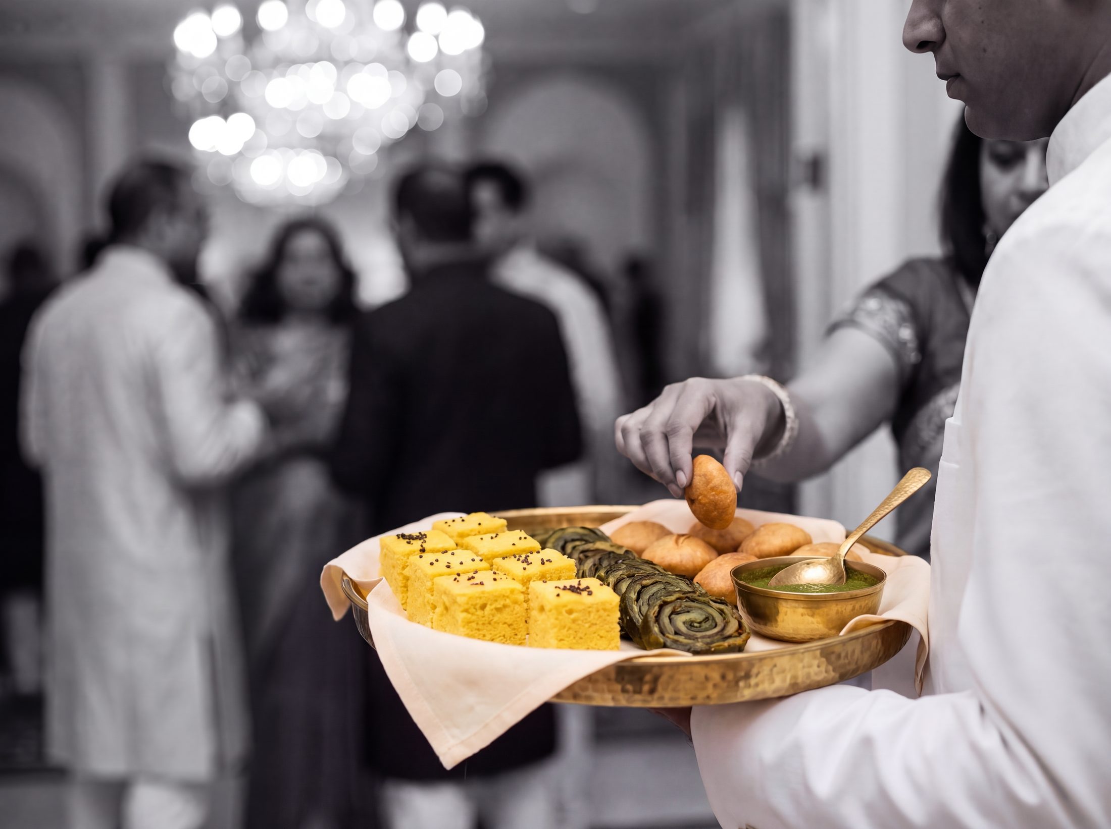 Server passing a brass tray of khaman dhokla, patra and lilva kachori at a wedding cocktail hour, guests in black and white