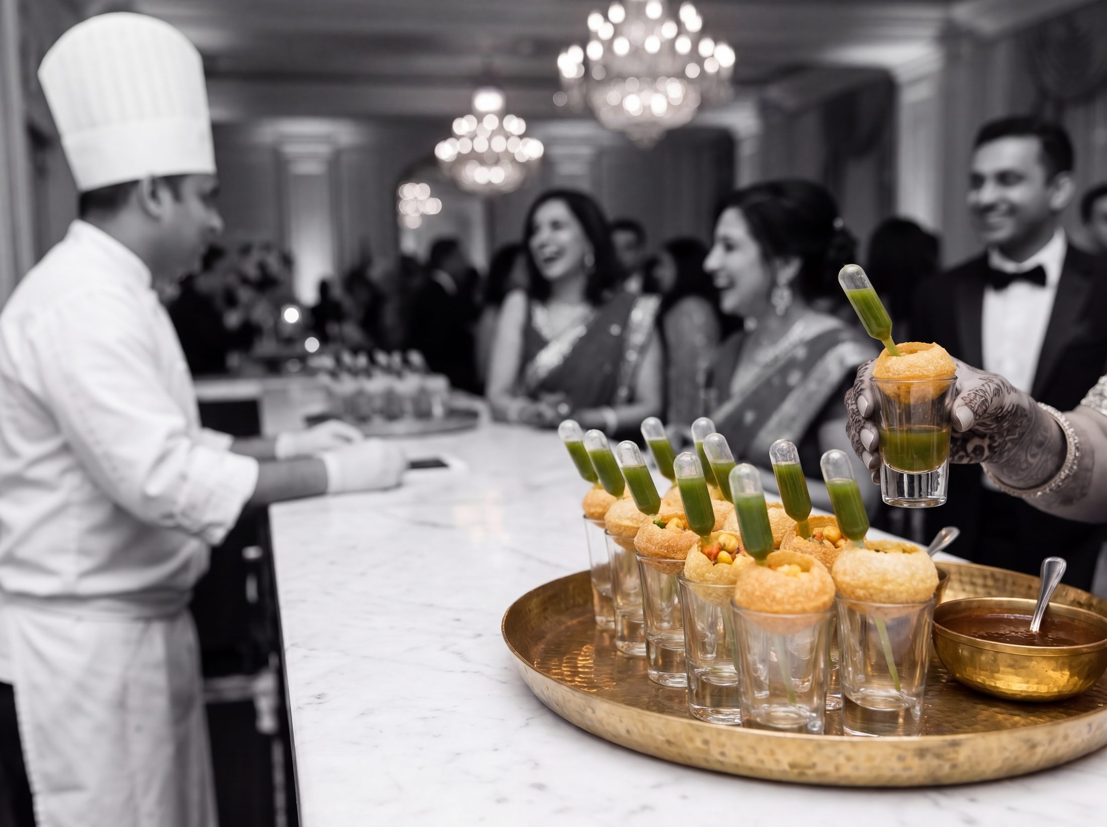 Guest lifting a pani puri shot glass with a pipette of green pani at a live chaat station, ballroom in black and white