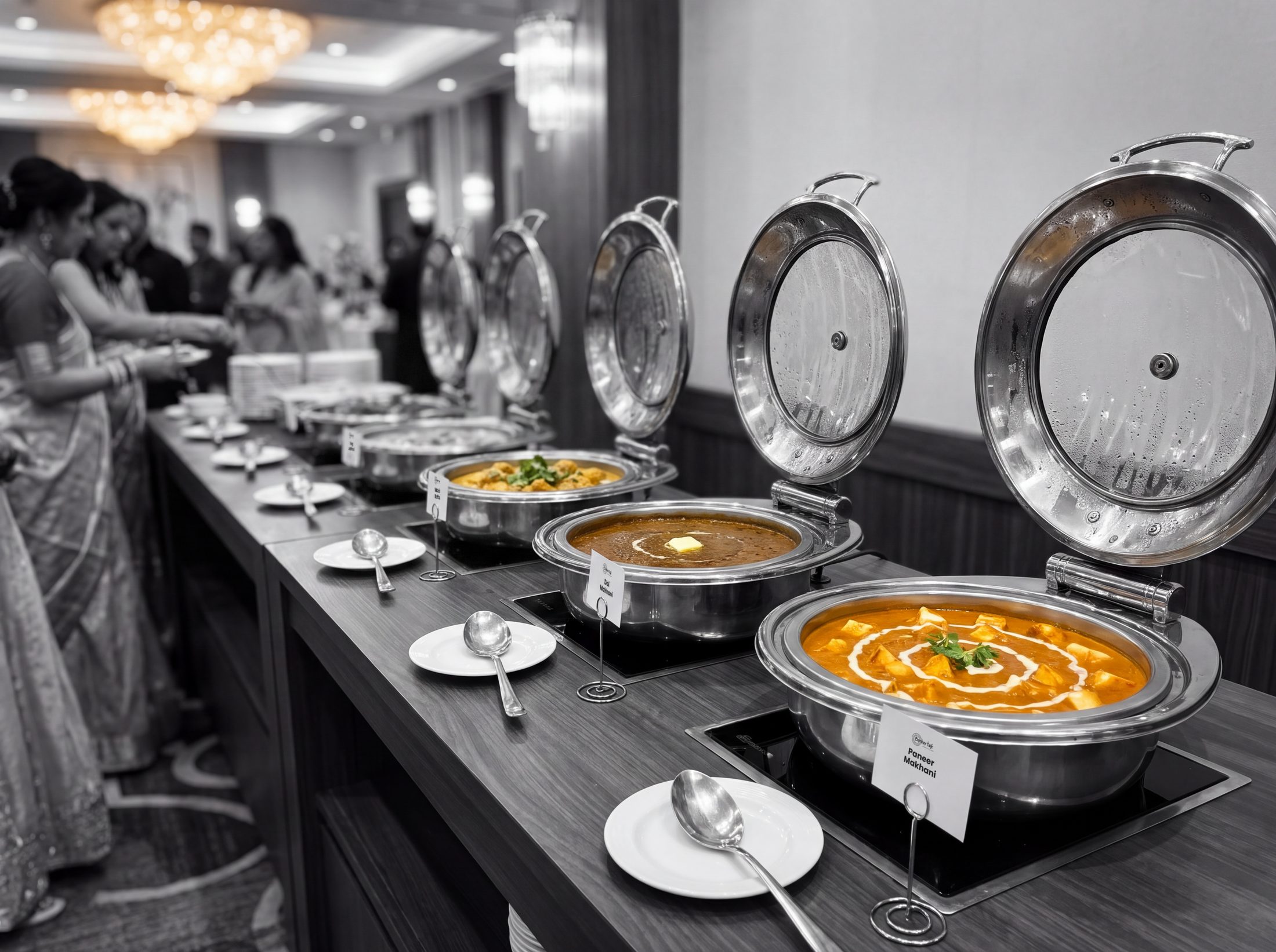 Paneer makhani, dal makhani and malai kofta in round induction chafers with glass lids on a wedding buffet, guests in black and white