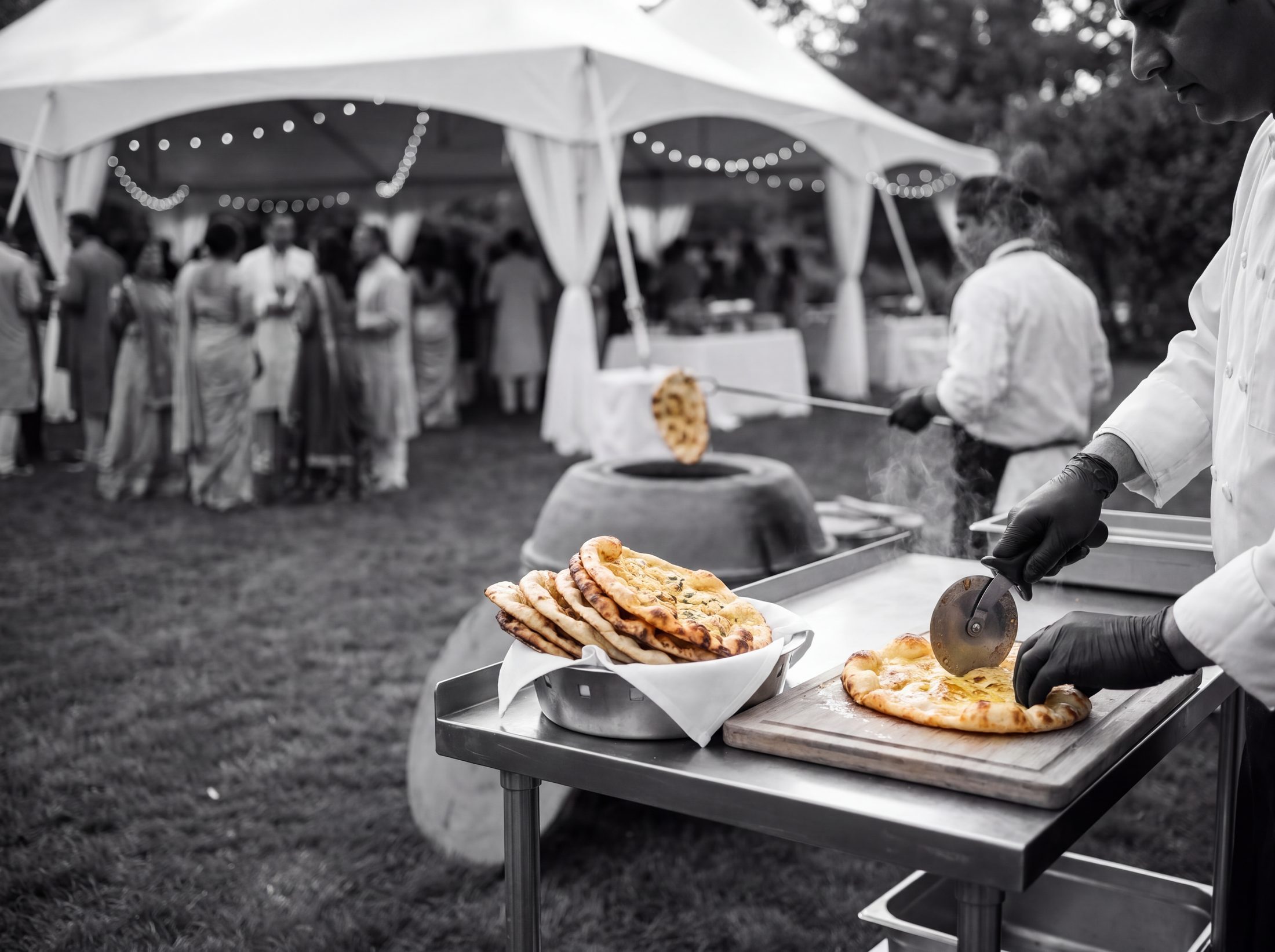 Chef slicing fresh butter naan at a prep table beside an outdoor tandoor at a garden wedding, in black and white
