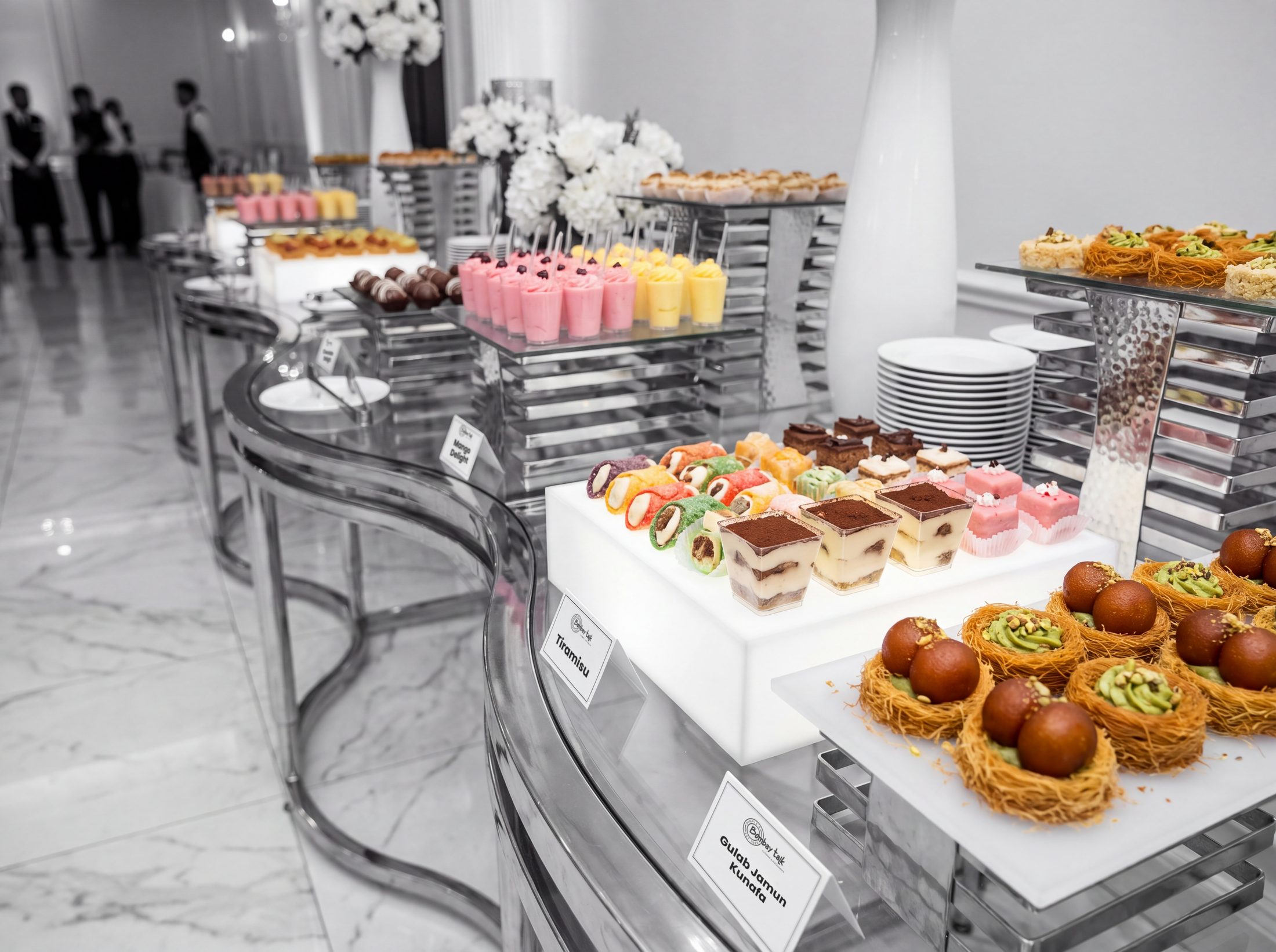 Gulab jamun on pistachio kunafa nests, tiramisu cups and mousse on chrome pedestals at a wedding dessert display, in black and white